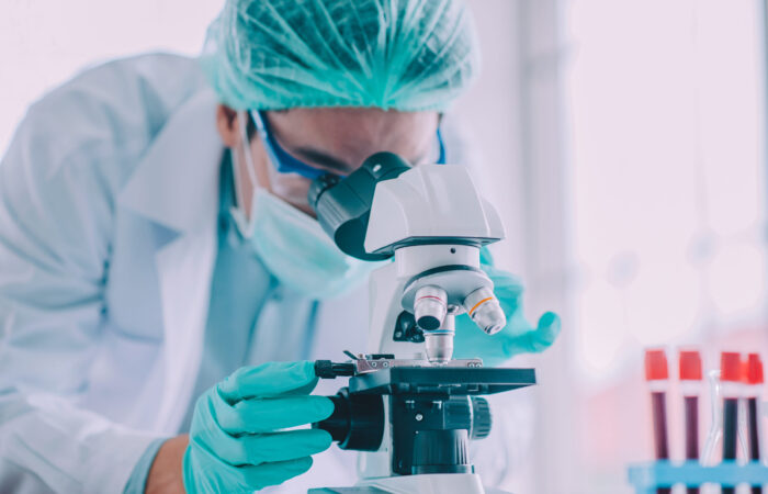 En medio de una cascada de tubos de ensayo que contienen un líquido rojo vibrante, un científico con bata de laboratorio, mascarilla verde y redecilla para el pelo observa a través de un microscopio. Los guantes azules garantizan un manejo cuidadoso mientras realizan meticulosamente una investigación para la publicación de salud Boletín Médico EuroEspes.