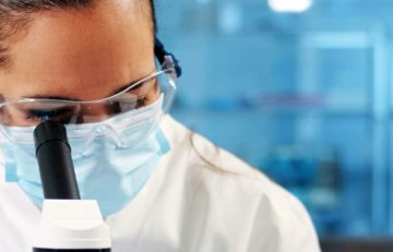 Un científico, con gafas de seguridad y mascarilla, examina minuciosamente muestras a través de un microscopio en el laboratorio de Salud de EuroEspes. El fondo está difuminado, destacando el material de laboratorio y envolviendo la escena en un suave tono azul.