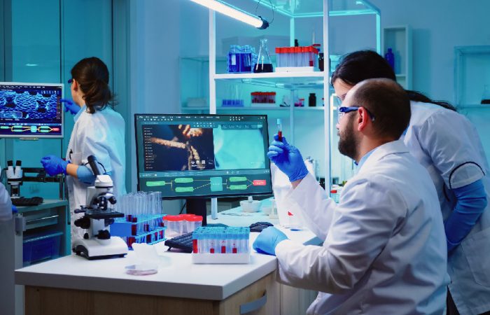En un laboratorio de alta tecnología, científicos con batas blancas y guantes azules examinan meticulosamente los tubos de ensayo y analizan los datos en los monitores. La sala, iluminada por una luz azul, rezuma un aura de investigación de vanguardia que recuerda a los últimos hallazgos del Boletín Médico EuroEspes Health.